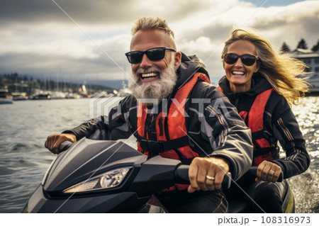Happy smiling elderly Caucasian couple in life vests riding jet ski on a lake or along sea coast. Active senior people having fun on water scooter. Healthy lifestyle for retired persons. Happy smiling elderly Caucasian couple in life vests riding jet ski on a lake or along sea coast. Active senior people having fun on water scooter. Healthy lifestyle for retired persons. 108316973