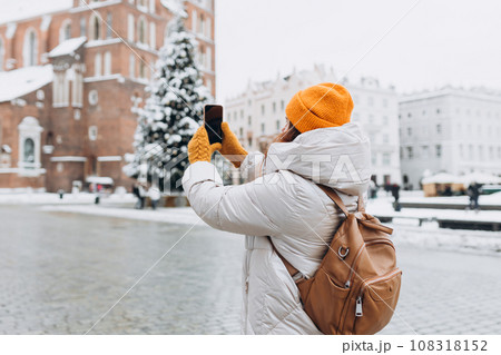 Attractive young female tourist is exploring new city. Redhead 30s woman in yellow hat resting on Market Square in Krakow. Traveling Europe in winter. St. Marys Basilica Attractive young female tourist is exploring new city. Redhead 30s woman in yellow hat resting on Market Square in Krakow. Traveling Europe in winter. St. Marys Basilica 108318152
