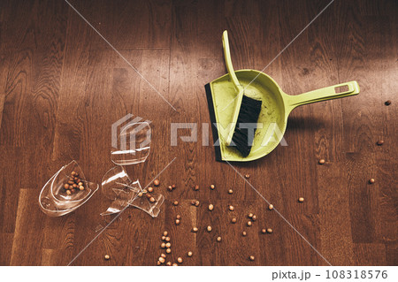 Top view of a brush with a dustpan and a broken transparent mug of compote with raisins. Shards of broken glass lie on the floor before cleaning with space to copy. High quality photo 108318576