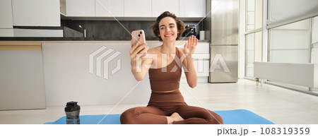 Smiling fitness girl, young athlete sits at home on yoga mat, waves hand at smartphone, joing online workout training session from home, says hello Smiling fitness girl, young athlete sits at home on yoga mat, waves hand at smartphone, joing online workout training session from home, says hello 108319359