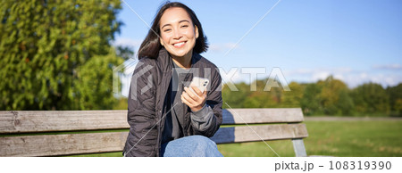 Cute young woman with smartphone in hands, sitting on bench and smiling, using mobile phone, waiting for someone in park Cute young woman with smartphone in hands, sitting on bench and smiling, using mobile phone, waiting for someone in park 108319390