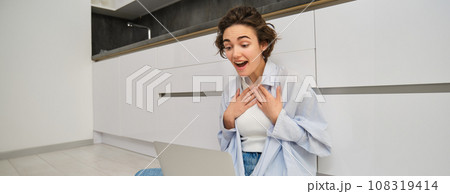 Portrait of girl sitting on floor with laptop, express surprise and excitement, hear amazing news while talking on video chat, having online conversation on her computer 108319414