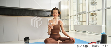 Sport app and wellbeing. Young woman, athlete sitting on floor at home with smartphone, doing yoga, using workout app on mobile phone, doing exercises at home Sport app and wellbeing. Young woman, athlete sitting on floor at home with smartphone, doing yoga, using workout app on mobile phone, doing exercises at home 108319417