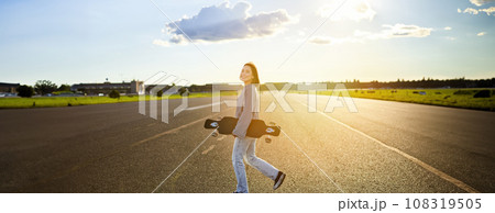 Asian girl with skateboard standing on road during sunset. Skater posing with her long board, cruiser deck during training Asian girl with skateboard standing on road during sunset. Skater posing with her long board, cruiser deck during training 108319505