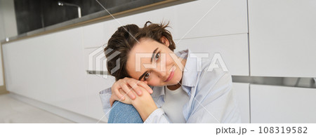 Close up portrait of beautiful, tender young woman smiling, looking at camera, sitting in cozy pose on kitchen floor, daydreaming Close up portrait of beautiful, tender young woman smiling, looking at camera, sitting in cozy pose on kitchen floor, daydreaming 108319582