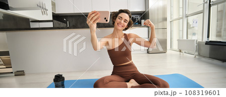 Fit and healthy woman fitness blogger, showing her muscles to smartphone camera, taking pictures in sportswear while doing yoga, workout at home and smiling 108319601