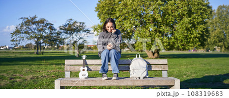 Excited girl looks at smartphone and celebrates, wins on mobile phone, sits with ukulele and backpack in park on bench on sunny day Excited girl looks at smartphone and celebrates, wins on mobile phone, sits with ukulele and backpack in park on bench on sunny day 108319683