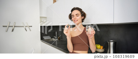 Dietary supplements and healthy lifestyle. Young woman taking vitamin C, D omega-3 with glass of water, standing in activewear, drinking after workout training in her kitchen Dietary supplements and healthy lifestyle. Young woman taking vitamin C, D omega-3 with glass of water, standing in activewear, drinking after workout training in her kitchen 108319689