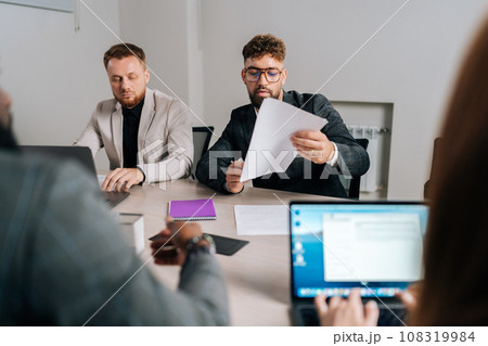 Serious young businessman reading contract document thinking considering risks with professional lawyers legal experts executive team, analyzing financial report sitting at office table. 108319984