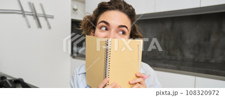 Portrait of cute brunette girl, looks away, hides her face behind planner, notebook, gets creative and writes in her journal Portrait of cute brunette girl, looks away, hides her face behind planner, notebook, gets creative and writes in her journal 108320972