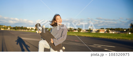 Funny asian girl enjoying skating, holding skateboard like guitar and shadow playing, having fun outdoors 108320975
