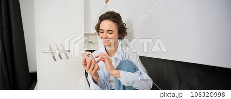 Smiling brunette girl sitting with smartphone at home, orders takeaway from mobile phone app, using online shopping application Smiling brunette girl sitting with smartphone at home, orders takeaway from mobile phone app, using online shopping application 108320998