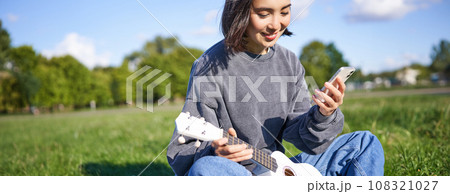 Technology and music. Smiling asian hipster girl sitting in park with smartphone and holding ukulele, playing instrument Technology and music. Smiling asian hipster girl sitting in park with smartphone and holding ukulele, playing instrument 108321027