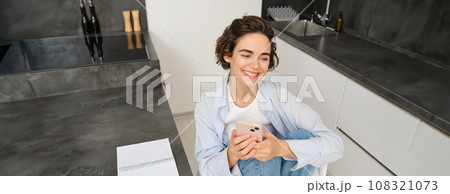 Young woman having break from work, sitting in kitchen and using smartphone, girl studies at home, sends a message on mobile phone 108321073