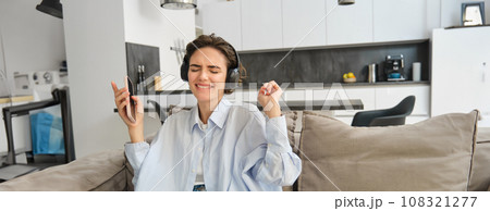 Portrait of beautiful, happy young woman, listening music in headphones, holding smartphone and dancing on sofa at home, enjoys spending time indoors alone Portrait of beautiful, happy young woman, listening music in headphones, holding smartphone and dancing on sofa at home, enjoys spending time indoors alone 108321277