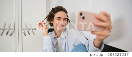 Smiling girl takes selfie on smartphone, posing for a photo on mobile phone app, sitting in her kitchen Smiling girl takes selfie on smartphone, posing for a photo on mobile phone app, sitting in her kitchen 108321337