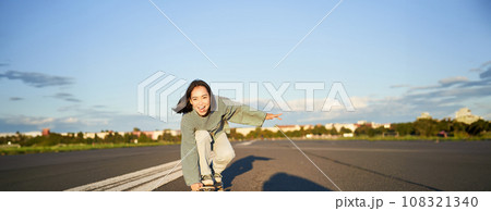 Skater girl riding on skateboard, standing on her longboard and laughing, riding cruiser on an empty street towards the sun 108321340