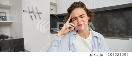 Portrait of woman with headache, sits in kitchen, touches head and grimaces, has painful migraine Portrait of woman with headache, sits in kitchen, touches head and grimaces, has painful migraine 108321395