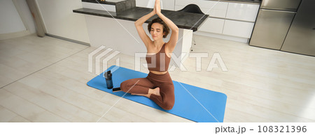 Portrait of fit and healthy woman in sportswear, doing yoga at home on fitness mat, making asana, relaxing and meditating indoors Portrait of fit and healthy woman in sportswear, doing yoga at home on fitness mat, making asana, relaxing and meditating indoors 108321396