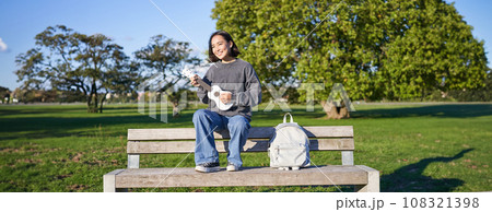 Portrait of young hipster girl sits on bench and plays ukulele, sings along, relaxing in park with her musical instrument 108321398