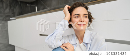 Portrait of young woman sits on floor, smiles and looks outside window, enjoys leisure at home, relaxes, has dreamy face expression 108321425