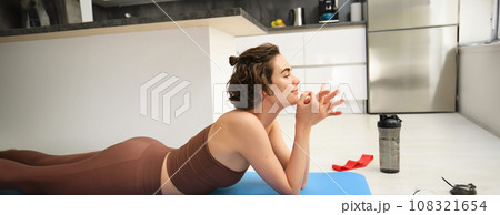 Sport and healthy lifestyle. Portrait of brunette sportswoman doing fitness in activewear, lying on rubber mat on floor, doing workout training in kitchen, drinking water from gym bottle Sport and healthy lifestyle. Portrait of brunette sportswoman doing fitness in activewear, lying on rubber mat on floor, doing workout training in kitchen, drinking water from gym bottle 108321654