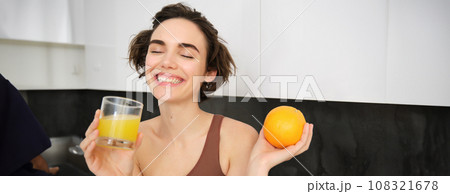 Sport and healthy lifestyle. Smiling happy young woman, holding an orange, drinking fresh juice and laughing, wearing activewear, standing in kitchen 108321678