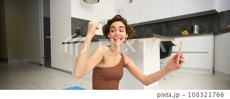 Young woman workout at home, rejoicing, using smartphone sport app, online video recordings from fitness instructor, celebrating, sitting on yoga mat on kitchen floor 108321766
