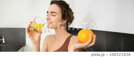 Healthy lifestyle and sport. Beautiful smiling woman, drinking fresh orange juice and holding fruit in her hand, enjoying her vitamin drink after workout at home, standing in kitchen Healthy lifestyle and sport. Beautiful smiling woman, drinking fresh orange juice and holding fruit in her hand, enjoying her vitamin drink after workout at home, standing in kitchen 108321836