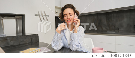 Portrait of young woman, 25 years old, holding smartphone, talking on mobile phone, sitting in kitchen at home 108321842
