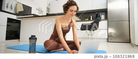Portrait of young female athlete, fitness instructor starting online gym class from her home, sitting on rubber yoga mat, typing on laptop 108321969