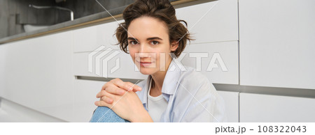 Close up portrait of young woman at home. Cute modern girl sitting on kitchen floor, smiling and looking at camera 108322043