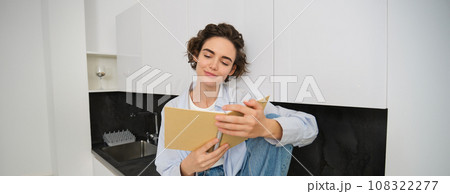 Portrait of smiling young woman reading journal, enjoys comfort at home, holding notebook, looking happy, relaxing indoors 108322277