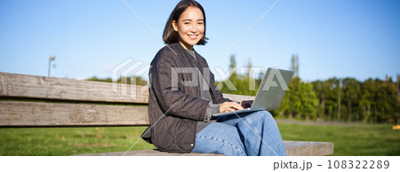 Portrait of smiling woman sitting with laptop, working on project or studying remotely, enjoying being in park 108322289