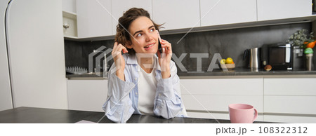 Smiling young woman talks on mobile phone, calls someone from home, sits in kitchen and has conversation 108322312