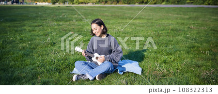 Happy brunette asian girl, student playing ukulele sitting on grass in park, relaxing, singing song, lifestyle concept Happy brunette asian girl, student playing ukulele sitting on grass in park, relaxing, singing song, lifestyle concept 108322313