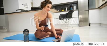 Portrait of young female athlete, yoga girl paying for online classes, purchasing remote training with gym instructor, sitting on her rubber mat with laptop and credit card 108322324