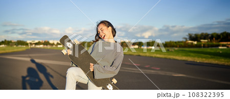 Funny asian girl enjoying skating, holding skateboard like guitar and shadow playing, having fun outdoors Funny asian girl enjoying skating, holding skateboard like guitar and shadow playing, having fun outdoors 108322395