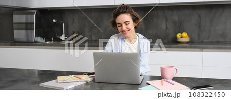 Image of young productive woman works from home, does distance learning, learns online course on her laptop in kitchen, writes down information, watches webinar, attends work meeting remotely Image of young productive woman works from home, does distance learning, learns online course on her laptop in kitchen, writes down information, watches webinar, attends work meeting remotely 108322450
