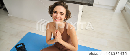 Peace, mindfulness and meditation. Young smiling sportswoman, fitness girl sits at home on floor and meditating, doing yoga, relaxing in lotus pose 108322456