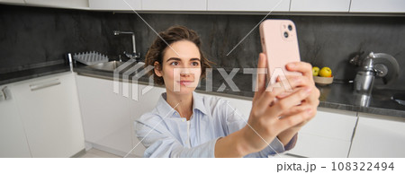 Portrait of beautiful young woman, taking selfie on mobile phone at home, poses for photo with smartphone Portrait of beautiful young woman, taking selfie on mobile phone at home, poses for photo with smartphone 108322494