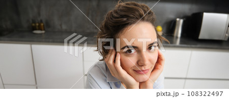 Close up portrait of young brunette woman, looks at camera and smiles, sits at home in kitchen 108322497