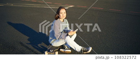 Sad asian teen girl sits on skateboard, looks upset, holds mobile phone and frowns Sad asian teen girl sits on skateboard, looks upset, holds mobile phone and frowns 108322572