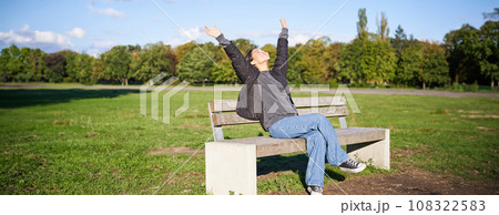 Portrait of happy asian girl feeling freedom and excitement, stretching hands while sitting on bench and smiling Portrait of happy asian girl feeling freedom and excitement, stretching hands while sitting on bench and smiling 108322583