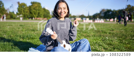 Portrait of excited asian girl, sitting with ukulele in park on green grass, looking amazed and surprised with good news 108322589
