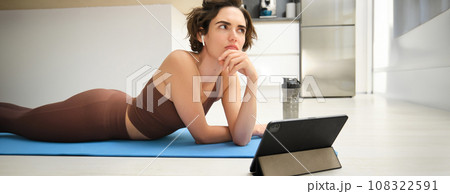 Portrait of fitness girl with thoughtful face, lying on rubber yoga mat at home, listening and watching sport workout video tutorial on tablet, thinking Portrait of fitness girl with thoughtful face, lying on rubber yoga mat at home, listening and watching sport workout video tutorial on tablet, thinking 108322591