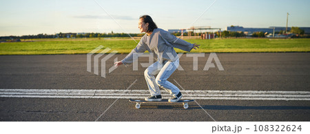 Happy skater girl riding her skateboard and having fun on empty street. Smiling woman enjoying cruiser ride on sunny road Happy skater girl riding her skateboard and having fun on empty street. Smiling woman enjoying cruiser ride on sunny road 108322624
