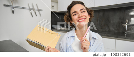 Portrait of smiling brunette girl, holds her planner, writes in notebook and looks happy, laughs, does homework, makes notes, sits at home in kitchen 108322691