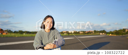 Vertical shot of asian woman sitting on skateboard on road, holding smartphone app. Skater girl skates on longboard, using mobile phone Vertical shot of asian woman sitting on skateboard on road, holding smartphone app. Skater girl skates on longboard, using mobile phone 108322722