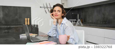 Beautiful brunette woman, looking outside window, working from home, drinking coffee in kitchen and suign laptop Beautiful brunette woman, looking outside window, working from home, drinking coffee in kitchen and suign laptop 108322768